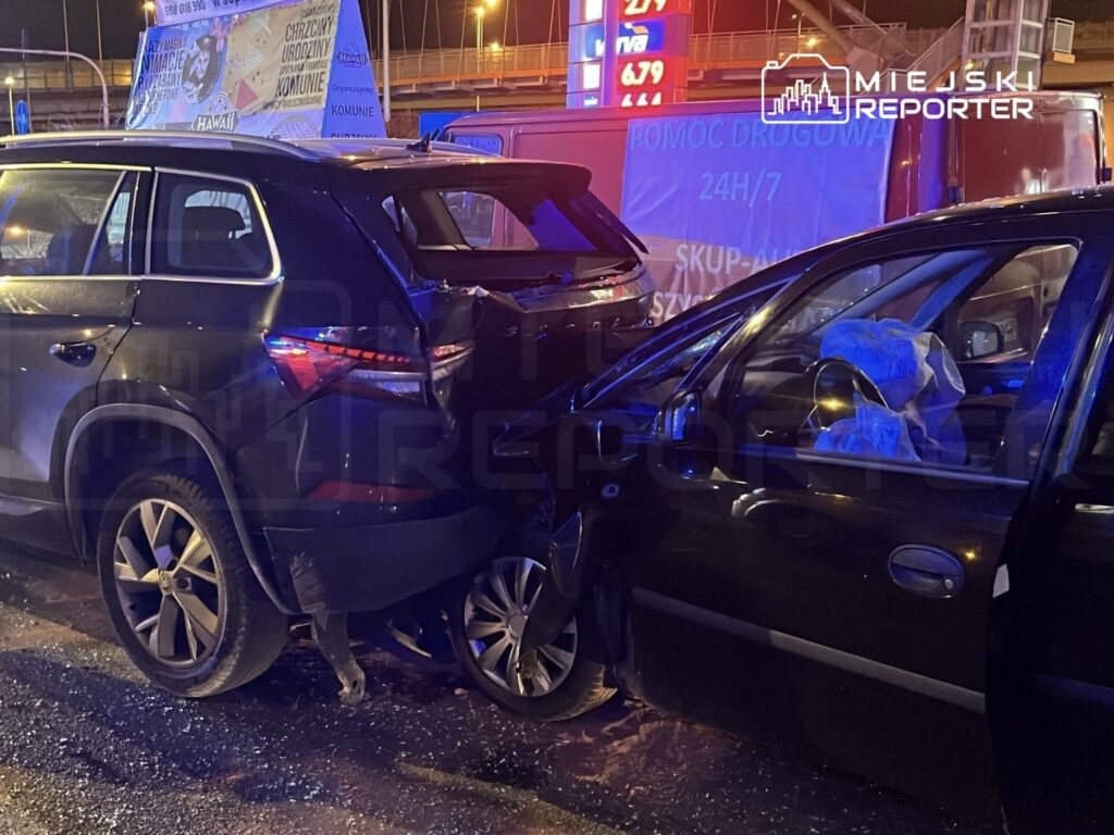 Dwa uszkodzone samochody osobowe zderzone na drodze, w tle widoczne neonowe reklamy i oświetlenie nocne.