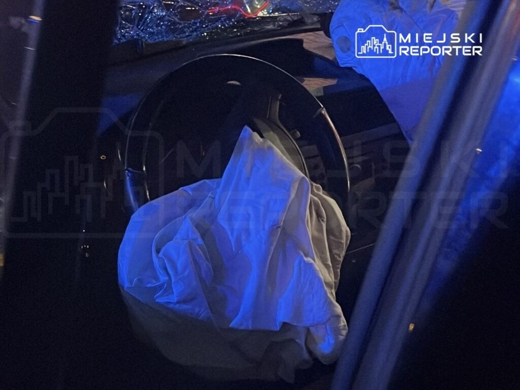 Biała tkanina leży na kierownicy samochodu, w tle widać rozbite szyby i niebieskie światła.