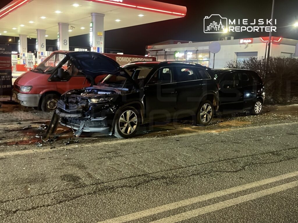 Dwa uszkodzone samochody osobowe stoją na poboczu obok stacji paliw, jeden z otwartą maską.