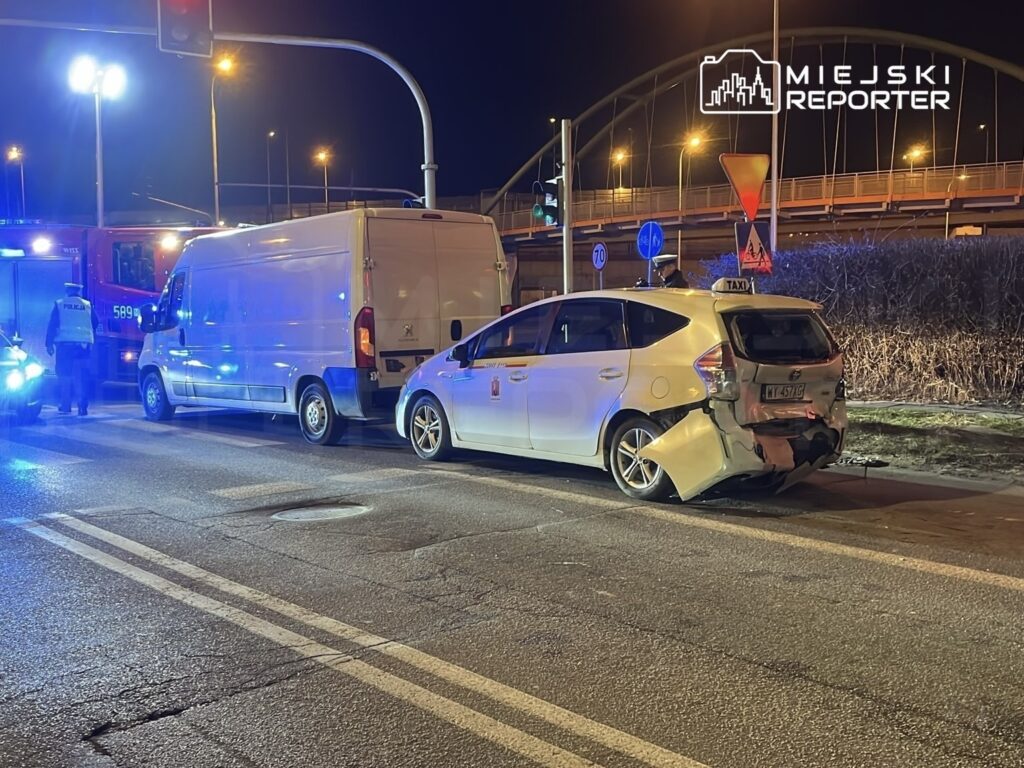 Na skrzyżowaniu ulic stoi biały samochód osobowy z uszkodzeniem tyłu, obok parkowany jest biały furgon. Policjant rozmawia z kierowcą.