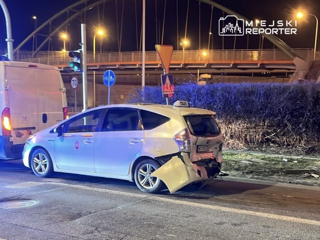 Uszkodzone taksówka z wgniecionym tyłem stoi na ulicy nocą, obok białego furgonu. W tle widać latarnie i znaki drogowe.