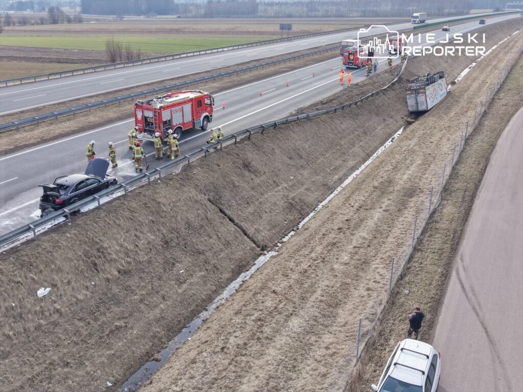 Grupa strażaków prowadzi akcję ratunkową przy uszkodzonym samochodzie na poboczu drogi, obok przewróconej ciężarówki.