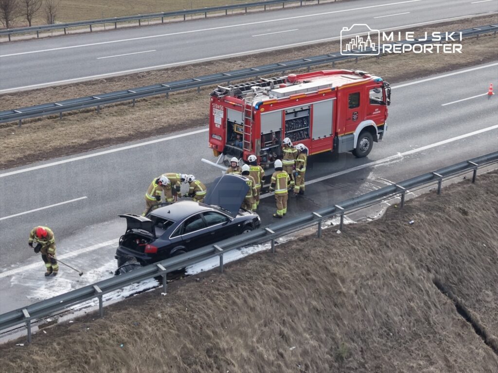 Strażacy w mundurach pracują nad czarnym samochodem osobowym z otwartą maską na poboczu drogi, obok stoi wóz strażacki.