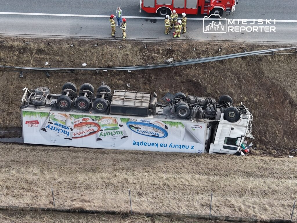 Cysterna przewożąca produkty spożywcze leży na boku w rowie przy drodze, obok grupy strażaków i ratowników.
