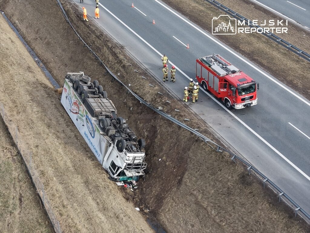 Cysterna przewrócona na stoku obok drogi, obok której pracują strażacy i pojazd gaśniczy.