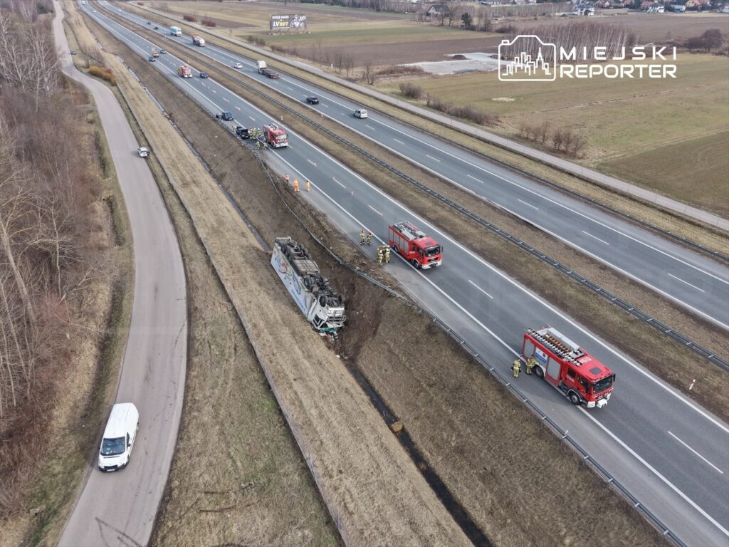 Na poboczu drogi leży przewrócony pojazd, obok pracują strażacy oraz pojazdy ratunkowe, w tle widoczna autostrada.