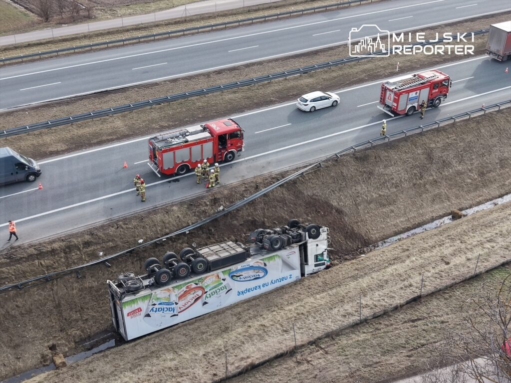 Cysterna przewrócona na poboczu drogi, obok pojazdy straży pożarnej i ratowników w akcji.