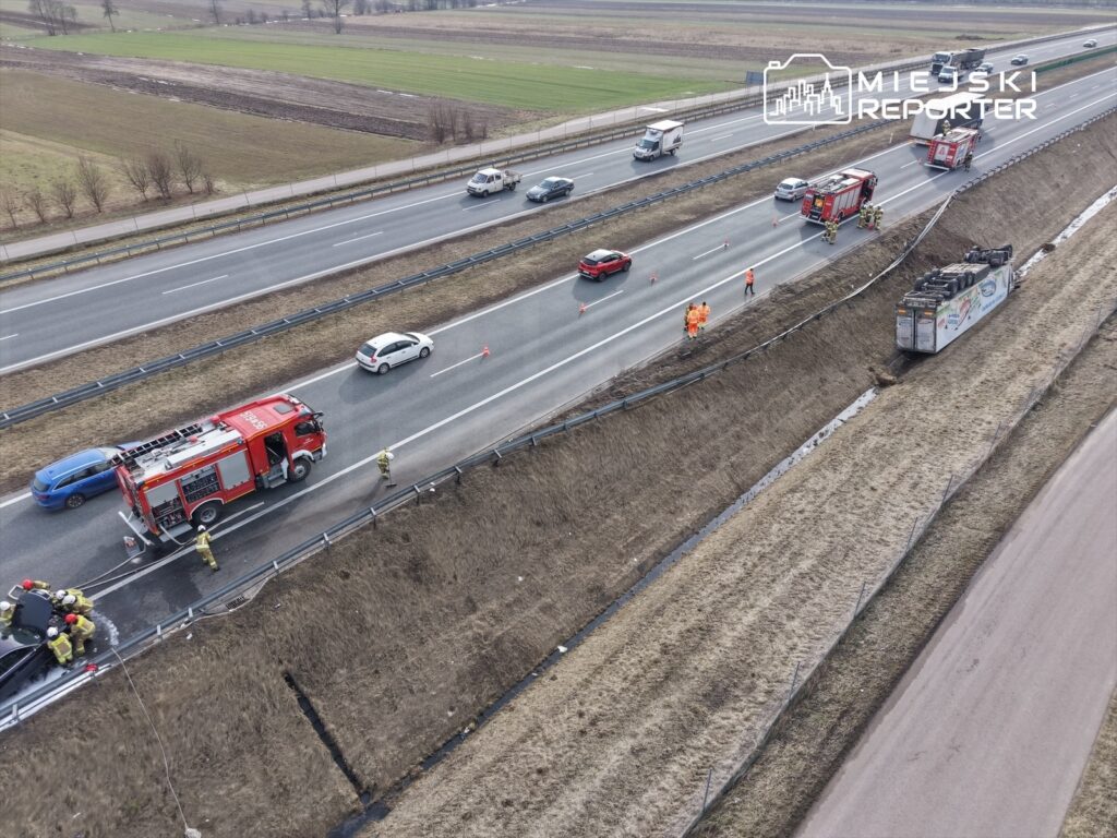Na drodze krajowej strażacy udzielają pomocy poszkodowanym po wypadku, obok przewrócony pojazd ciężarowy.