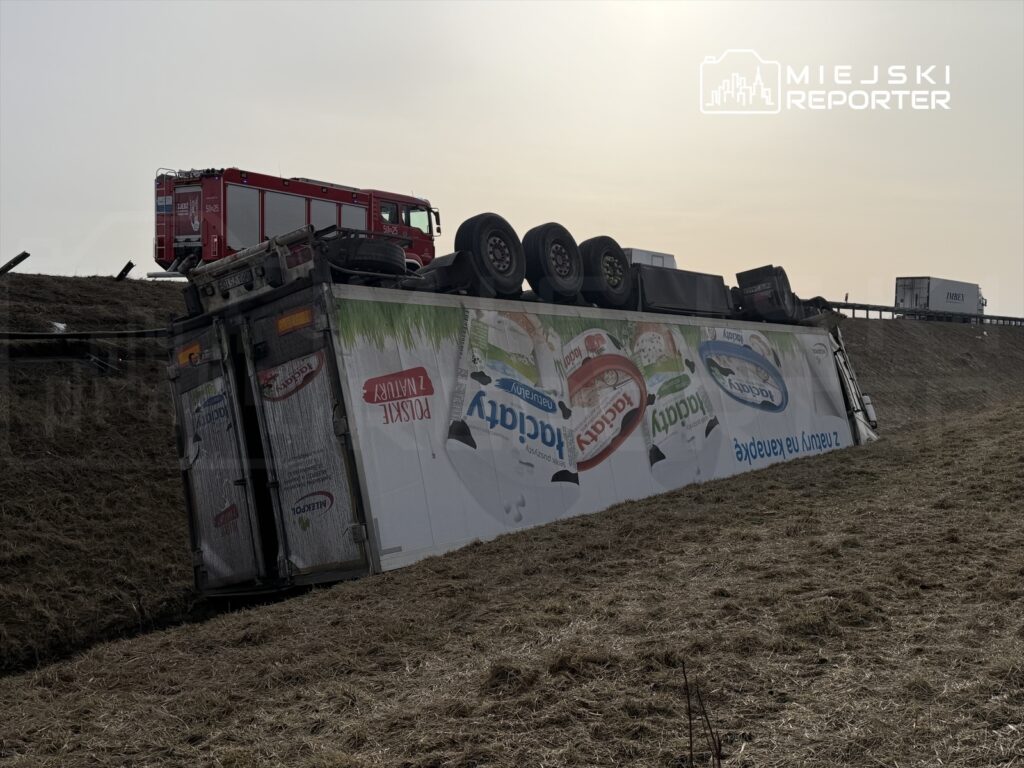 Cysterna przewożąca produkty mleczne leży na boku w rowie, obok stoi wóz strażacki.