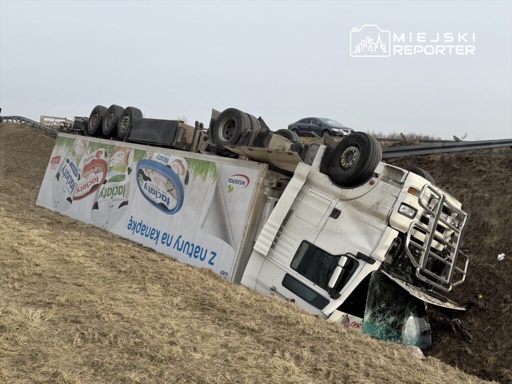 Wielka ciężarówka przewrócona na bok w rowie, z widoczną reklamą produktów mlecznych na boku. W tle widać inne pojazdy na drodze.