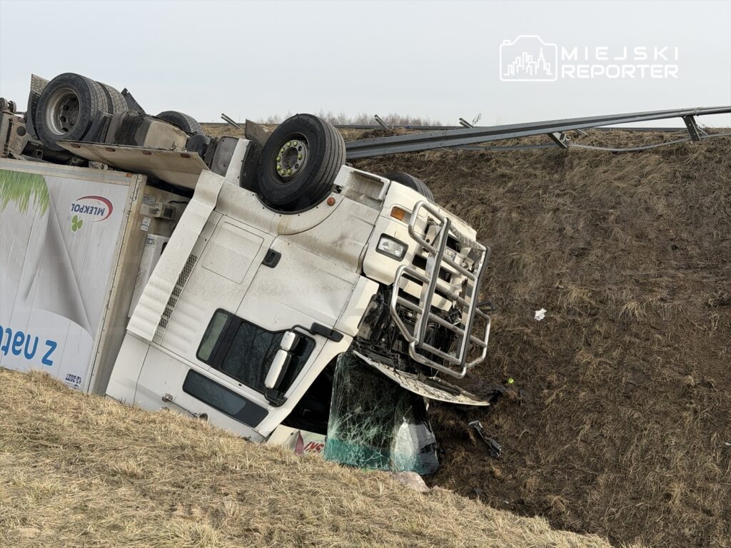 Przewrócony ciężarowy samochód na poboczu drogi, z uszkodzoną kabiną i wygiętym ogrodzeniem.