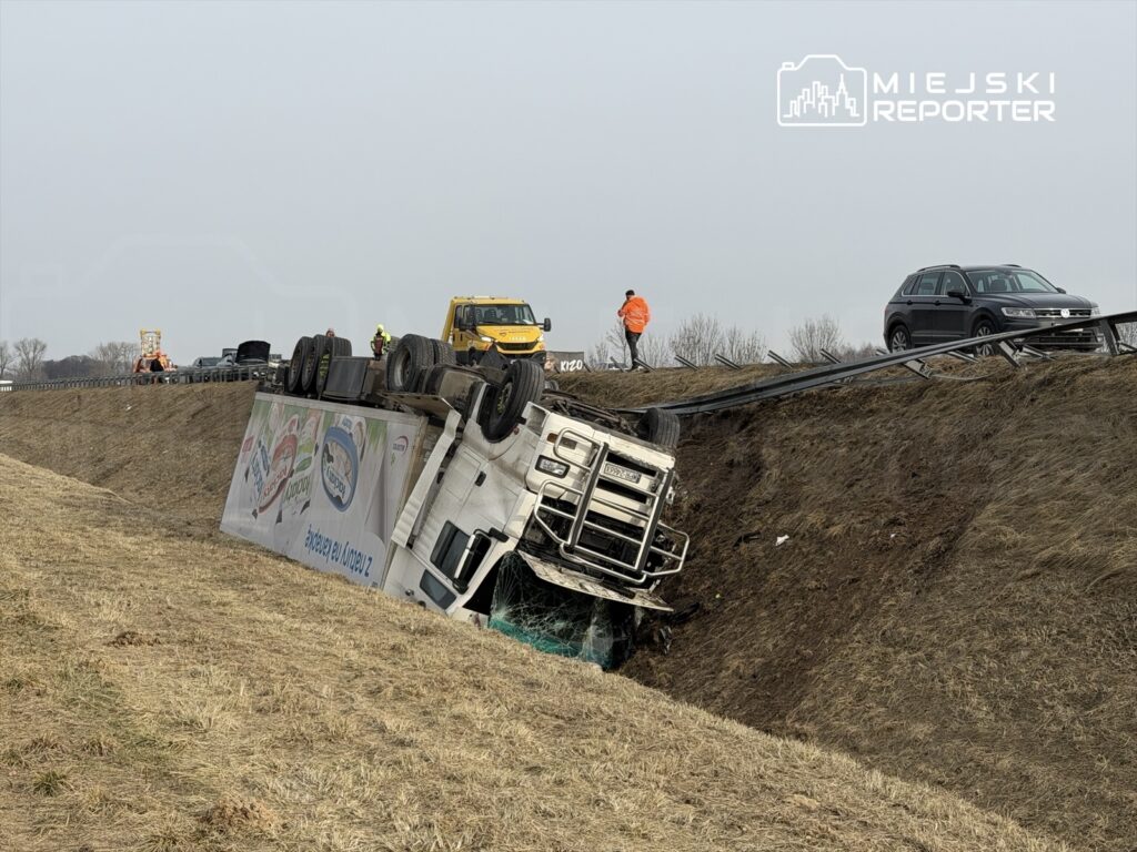 Ciężarówka leżąca na boku w rowie, przy drodze, obok pracujący ratownicy i pojazdy pomocy drogowej.