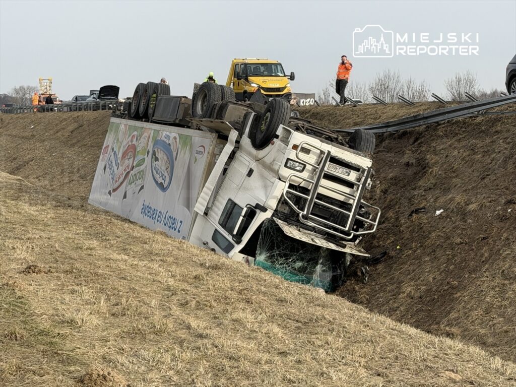Ciężarówka przewrócona na bok w rowie obok drogi, z widoczną zniszczoną szybą i reklamą na burcie. W tle pracują służby ratunkowe.