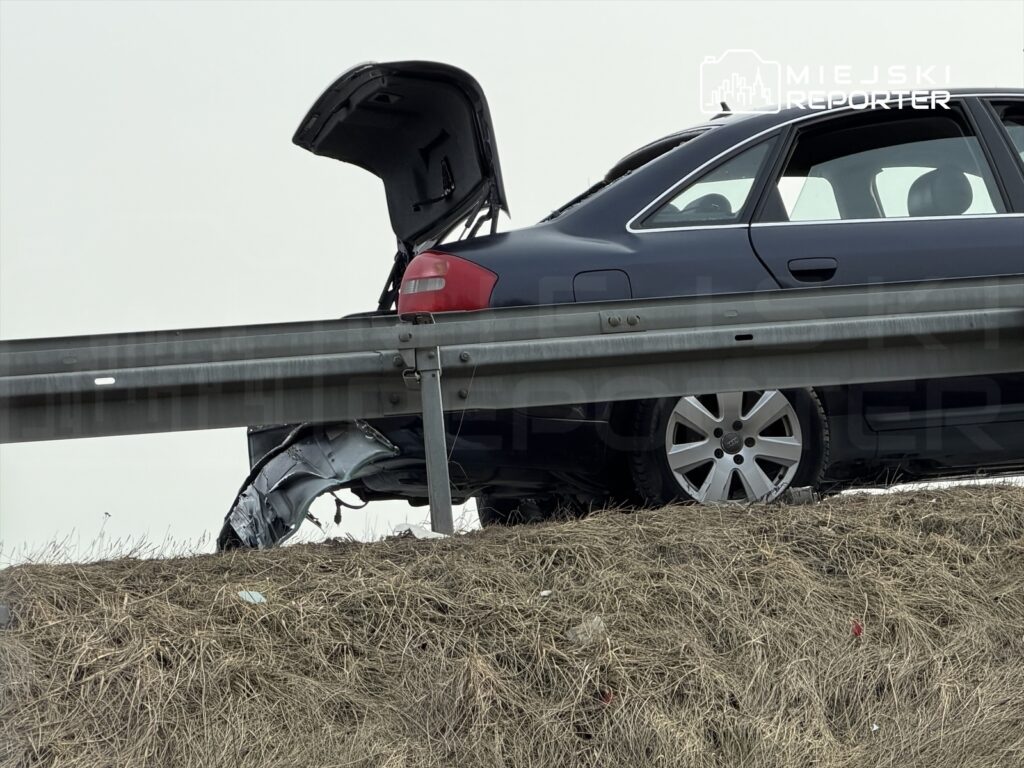 Uszkodzony samochód osobowy z otwartym bagażnikiem stoi na poboczu drogi, obok barierki ochronnej.