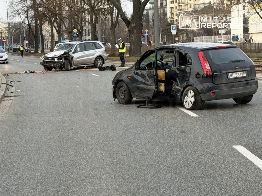 Dwa uszkodzone samochody po kolizji na ulicy, obok policjanci zabezpieczający miejsce zdarzenia.