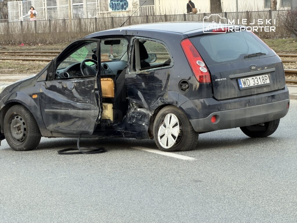 Czarny samochód osobowy z uszkodzeniami stoi na ulicy, z otwartymi drzwiami z lewej strony.
