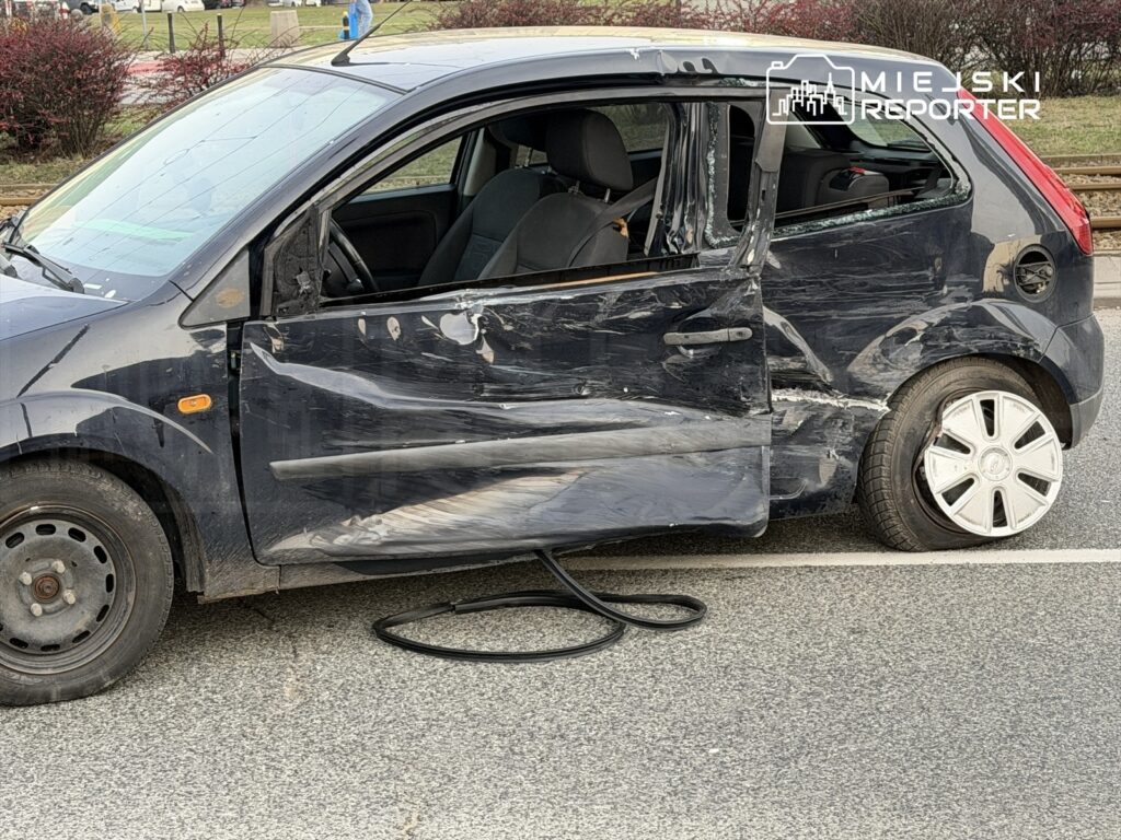 Uszkodzone czarne auto osobowe z otwartymi drzwiami, uszkodzoną karoserią i jednym płaskim kołem na ulicy.