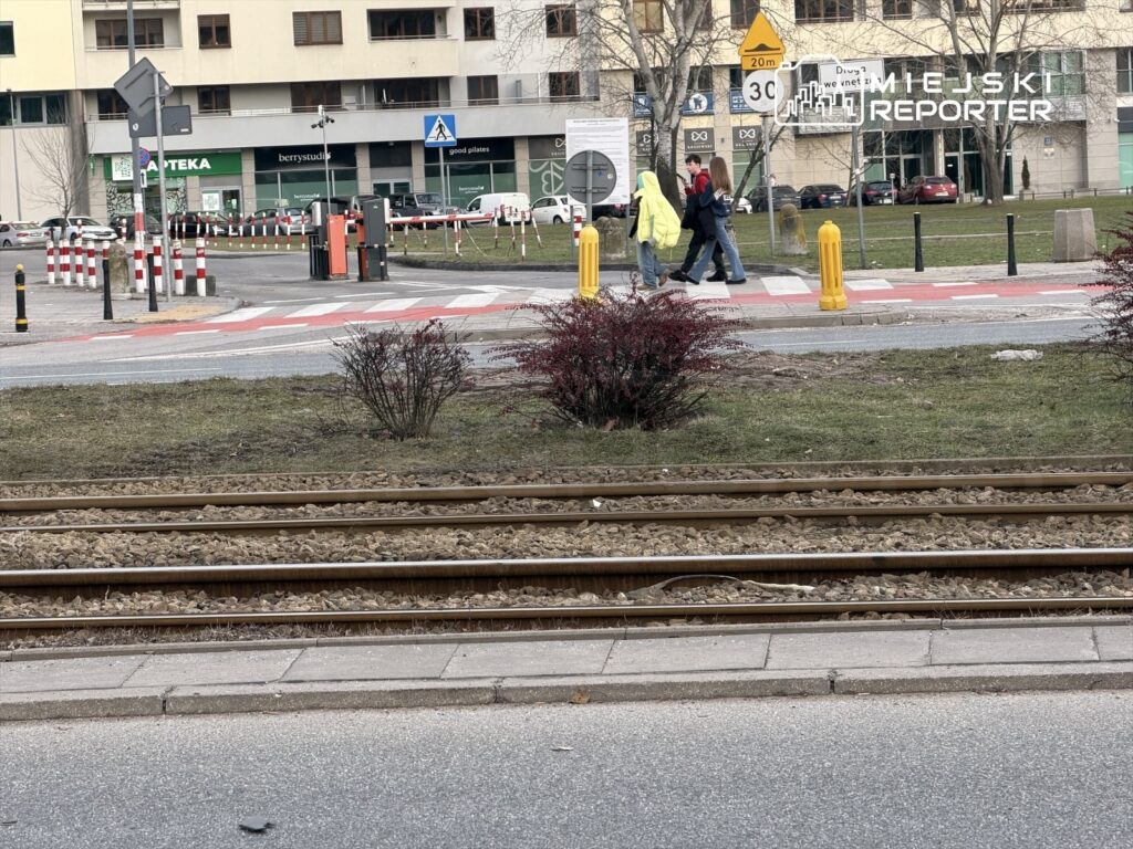 Grupa przechodniów w jasnych kurtkach przechodzi przez ulicę obok torów tramwajowych w miejskim otoczeniu.