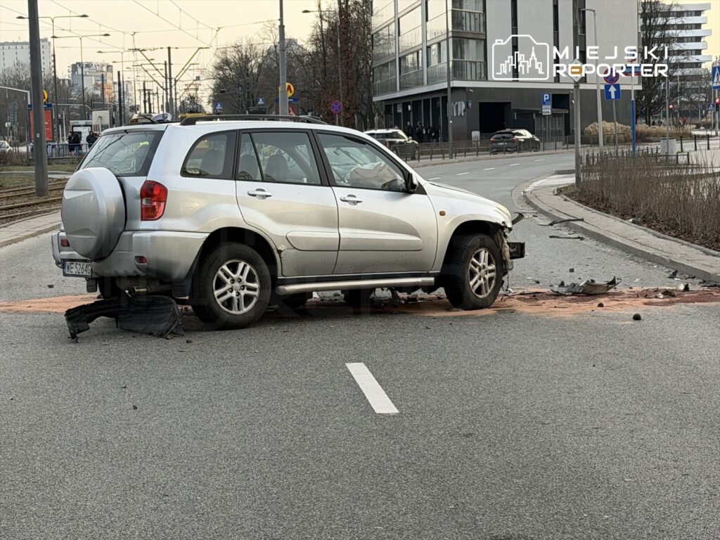 Uszkodzony srebrny SUV stoi na jezdni, obok leży fragment zderzaka oraz plama płynów.