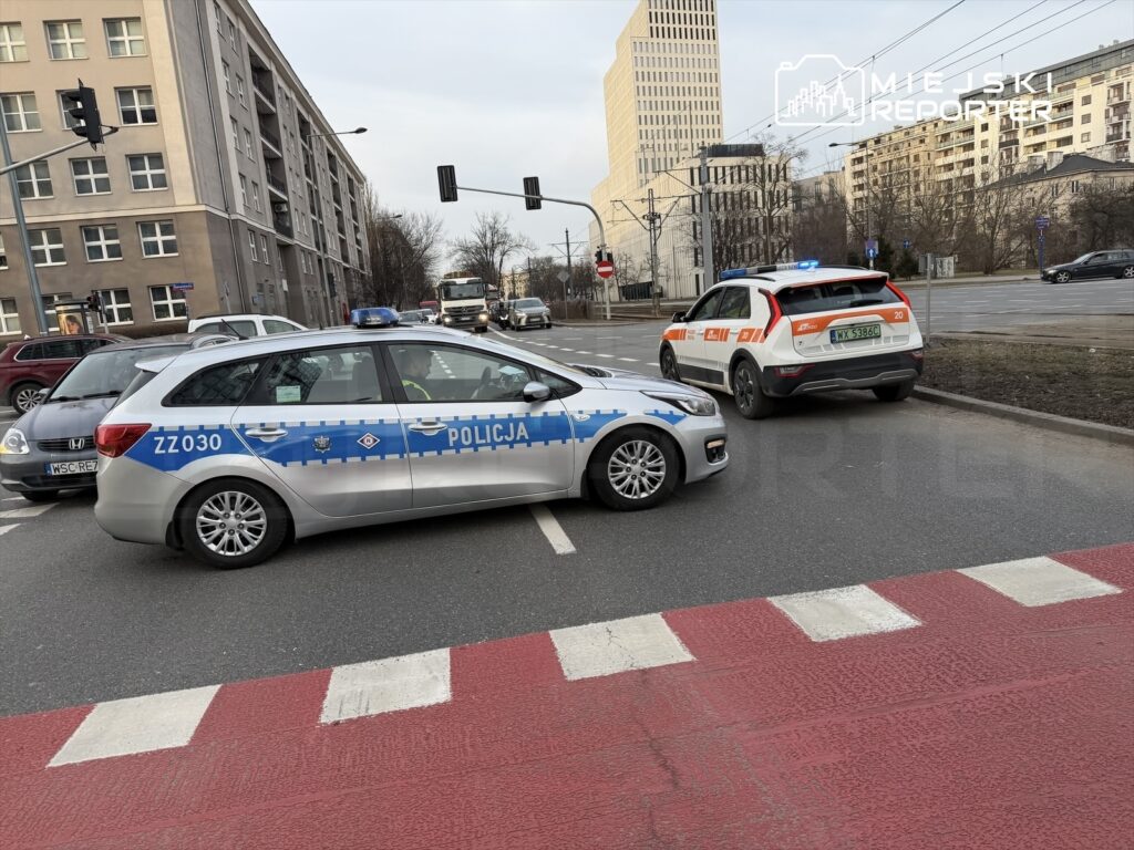 Policyjny radiowóz z napisem "POLICJA" blokujący ruch na skrzyżowaniu, obok pojazd straży miejskiej.