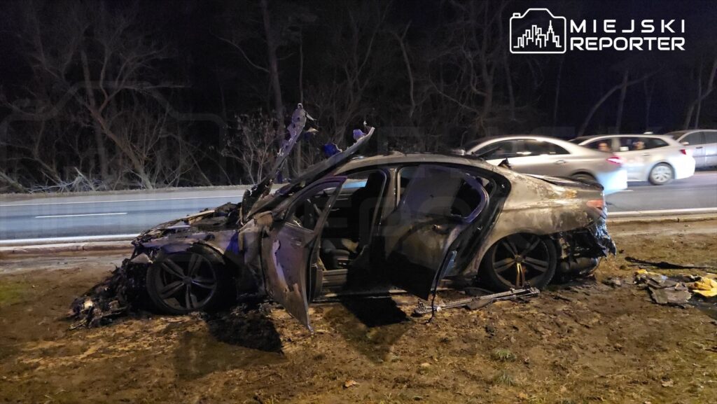 Spalony samochód osobowy stoi na poboczu drogi, obok ruchliwej jezdni, w otoczeniu nagich drzew.