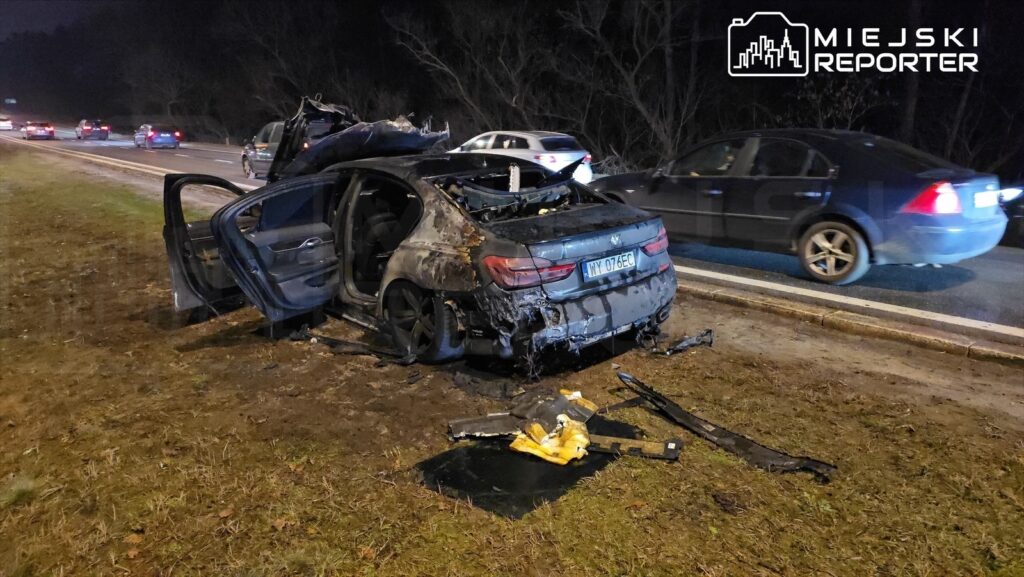 Zniszczony samochód osobowy stoi na poboczu drogi, obok przejeżdżające pojazdy w nocy.