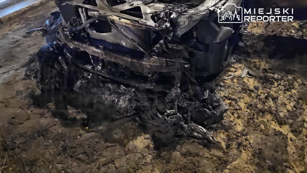 Spalone wraki samochodu leżące na ziemi, otoczone zniszczoną nawierzchnią drogi.