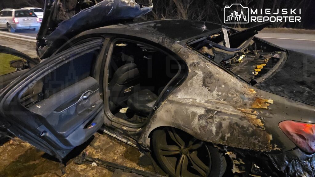 Spalony samochód osobowy z otwartymi drzwiami stoi na poboczu drogi, z uszkodzoną karoserią i wnętrzem.