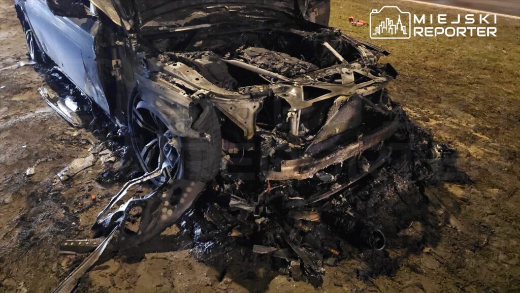 Spalony wrak samochodu osobowego leżący na poboczu drogi, obok widoczny rozrzucony popiół i resztki.