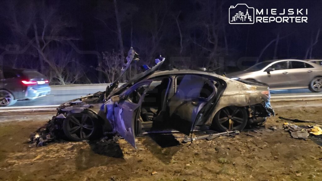 Spalony samochód osobowy stoi na poboczu drogi, obok przejeżdżające pojazdy. W tle widoczne drzewa i ciemne niebo.