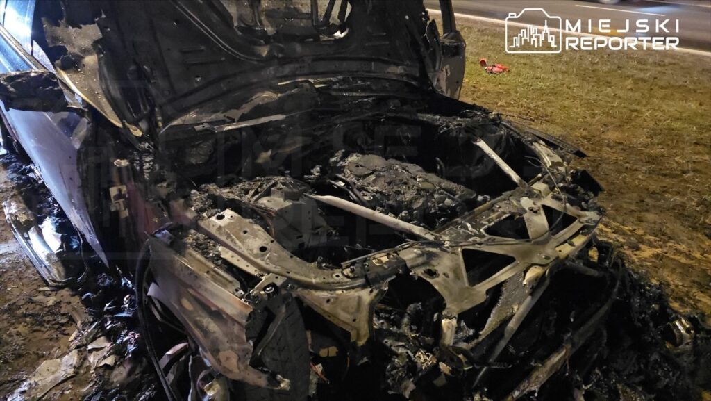 Spalony wrak samochodu osobowego leżący na poboczu drogi, z widocznymi uszkodzeniami silnika i nadwozia.