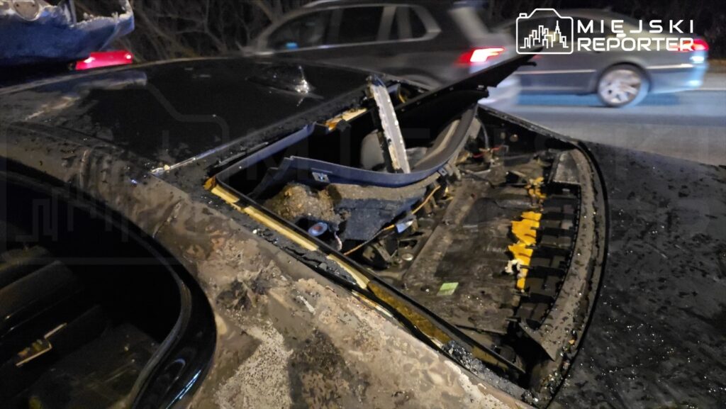 Uszkodzone auto z otwartą pokrywą bagażnika, z widocznymi elementami wnętrza i śladami pożaru, na drodze nocą.