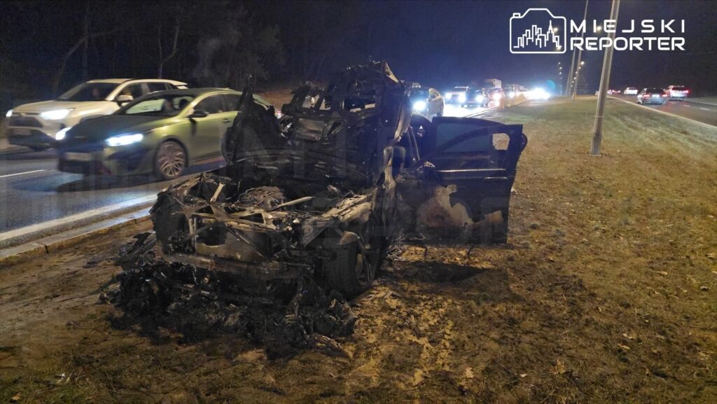 Spalony wrak samochodu osobowego stoi na poboczu drogi, obok przejeżdżające pojazdy w nocy.