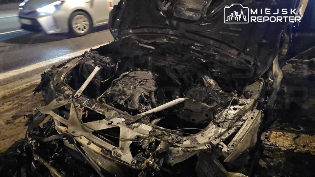 Spalony wrak samochodu osobowego leży na poboczu drogi, obok przejeżdżających pojazdów.