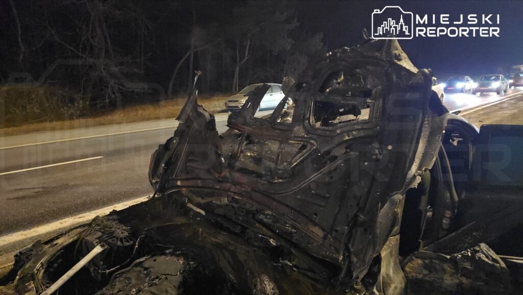 Spalony pojazd osobowy leży na poboczu drogi, obok przejeżdżające samochody. W tle widoczne drzewa.