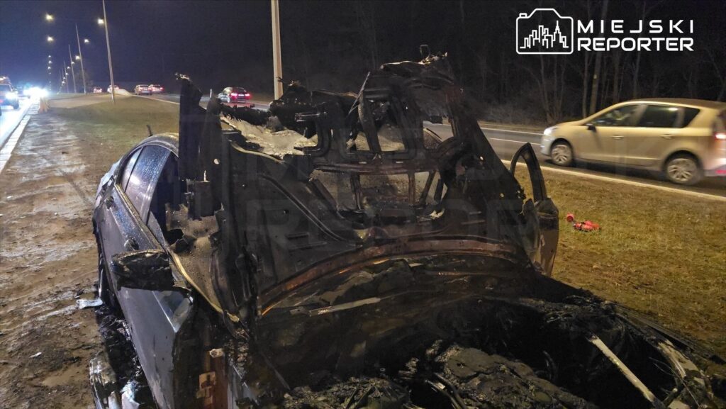 Spalony wrak samochodu osobowego stoi na poboczu drogi, obok przejeżdżające pojazdy w nocy.