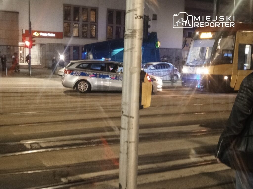 Policyjny radiowóz zatrzymany na skrzyżowaniu z tramwajem, w tle widoczny budynek Poczty Polskiej.