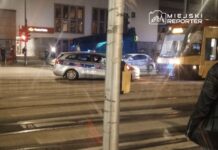 Policyjny radiowóz zatrzymany na skrzyżowaniu z tramwajem, w tle widoczny budynek Poczty Polskiej.