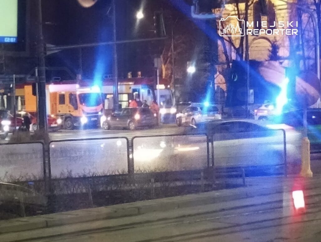 Karetka pogotowia oraz radiowozy policyjne na ruchliwej ulicy nocą, otoczone przez zaparkowane samochody.