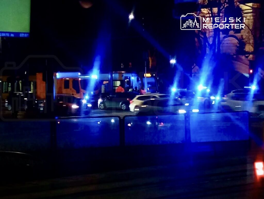 Na skrzyżowaniu zatrzymane pojazdy oraz radiowozy z włączonymi sygnałami świetlnymi. W tle widoczny tramwaj.