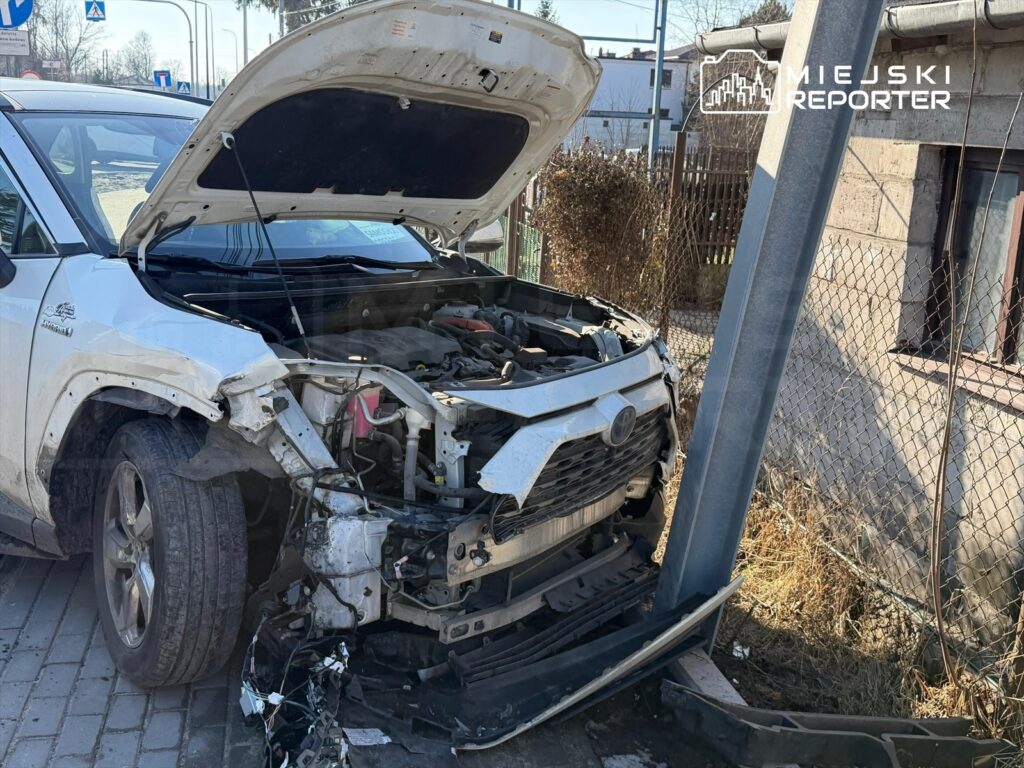 Uszkodzony samochód osobowy z otwartą maską stoi obok metalowego słupa, w tle widoczny płot i budynek.