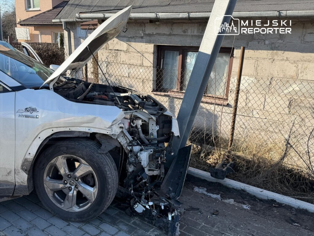 Uszkodzony samochód osobowy z otwartą maską uderzył w słup, stojąc na wybrukowanej drodze obok budynku mieszkalnego.