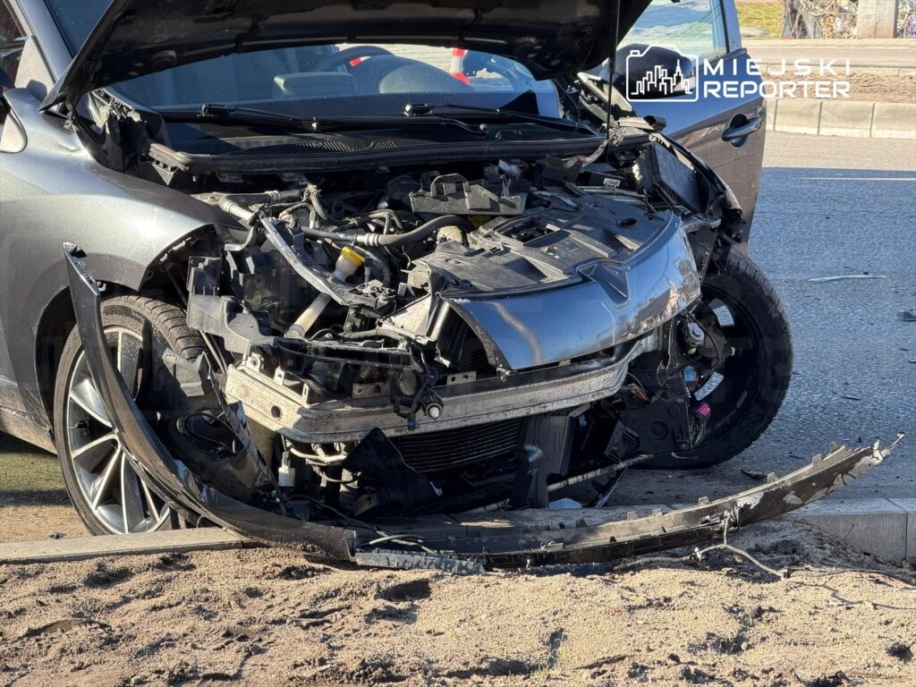 Zniszczony samochód osobowy z otwartą maską, uszkodzona przednia część i odłamana osłona zderzaka, na drodze.