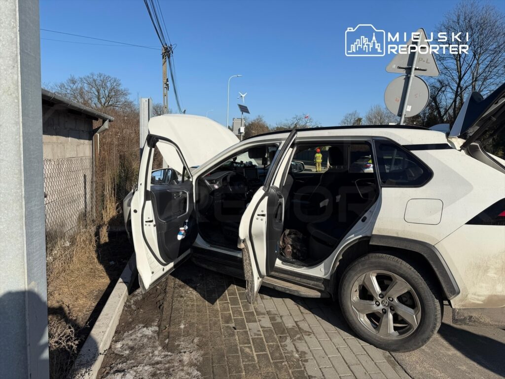 Biały samochód osobowy z otwartymi drzwiami zaparkowany przy drodze, obok widoczny znak drogowy.