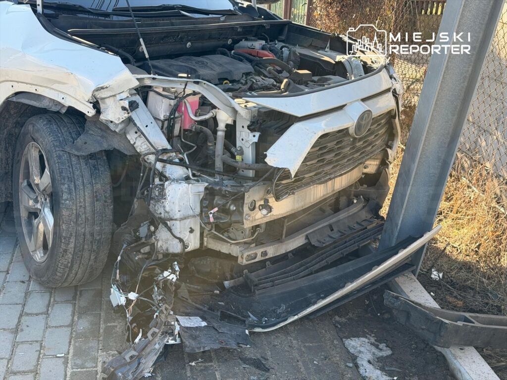 Uszkodzony samochód osobowy z wgniecionym przodem stoi obok metalowego słupa, na powierzchni bruku widoczne zniszczenia.