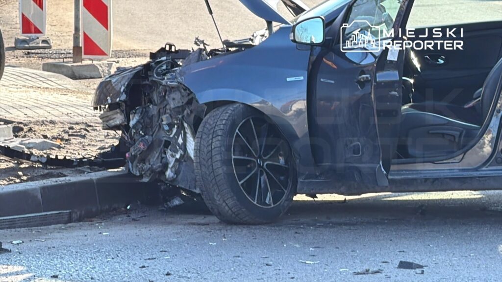 Zniszczony samochód osobowy stoi na poboczu drogi, z wyraźnymi uszkodzeniami przodu i otwartymi drzwiami.