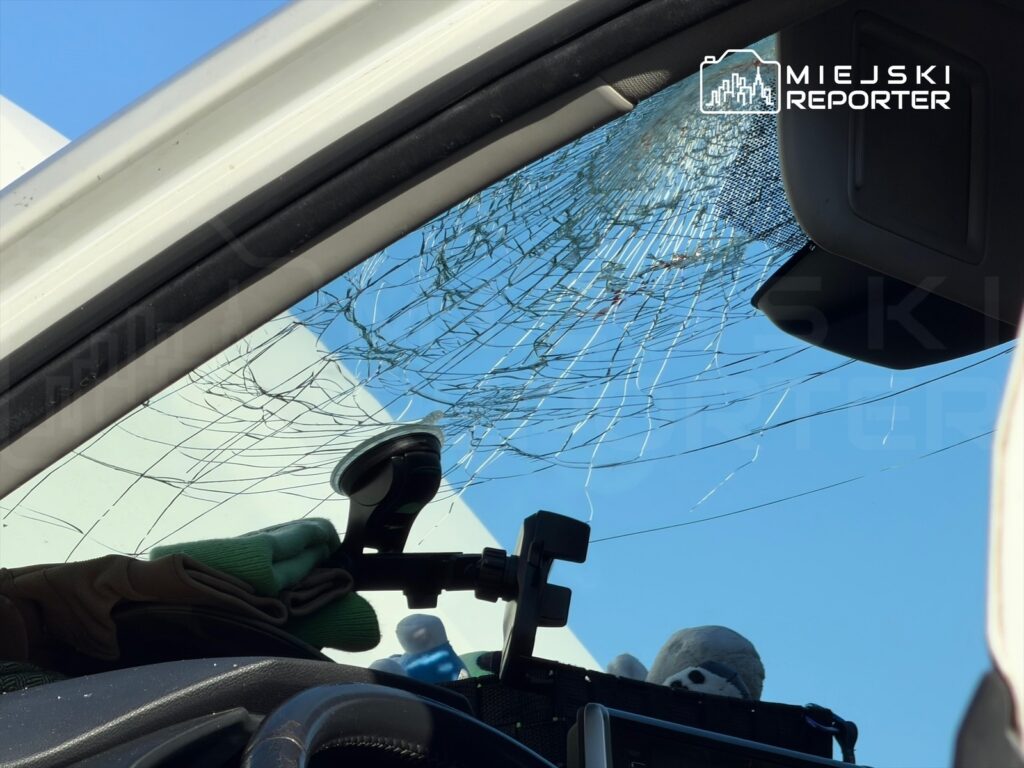 Pęknięta szyba samochodowa z widokiem na błękitne niebo oraz rękawice ochronne leżące na desce rozdzielczej.