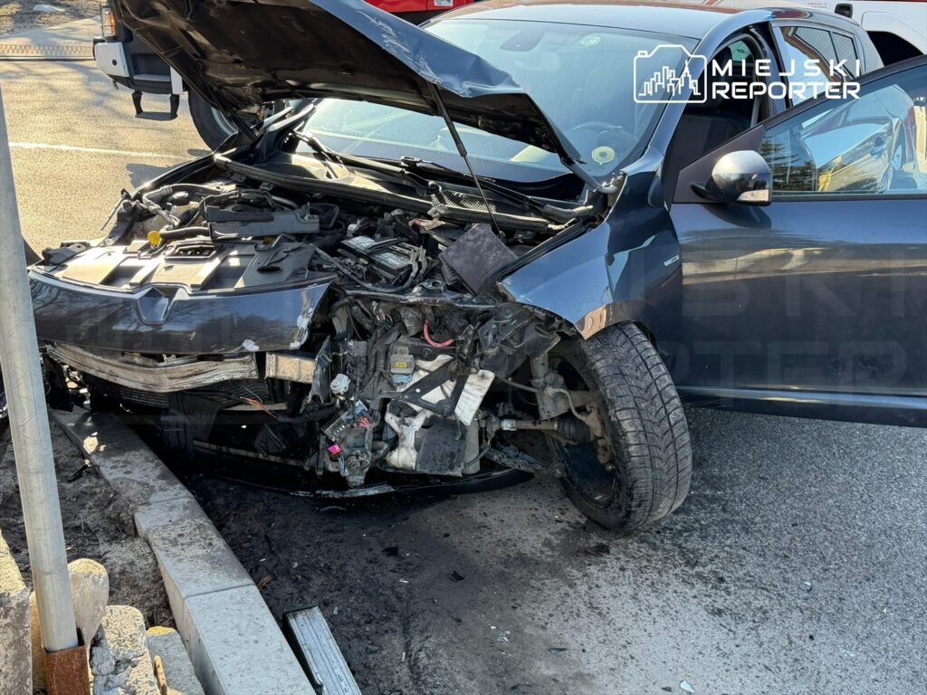 Uszkodzony samochód osobowy z otwartą maską, oparty o słup, na poboczu drogi.