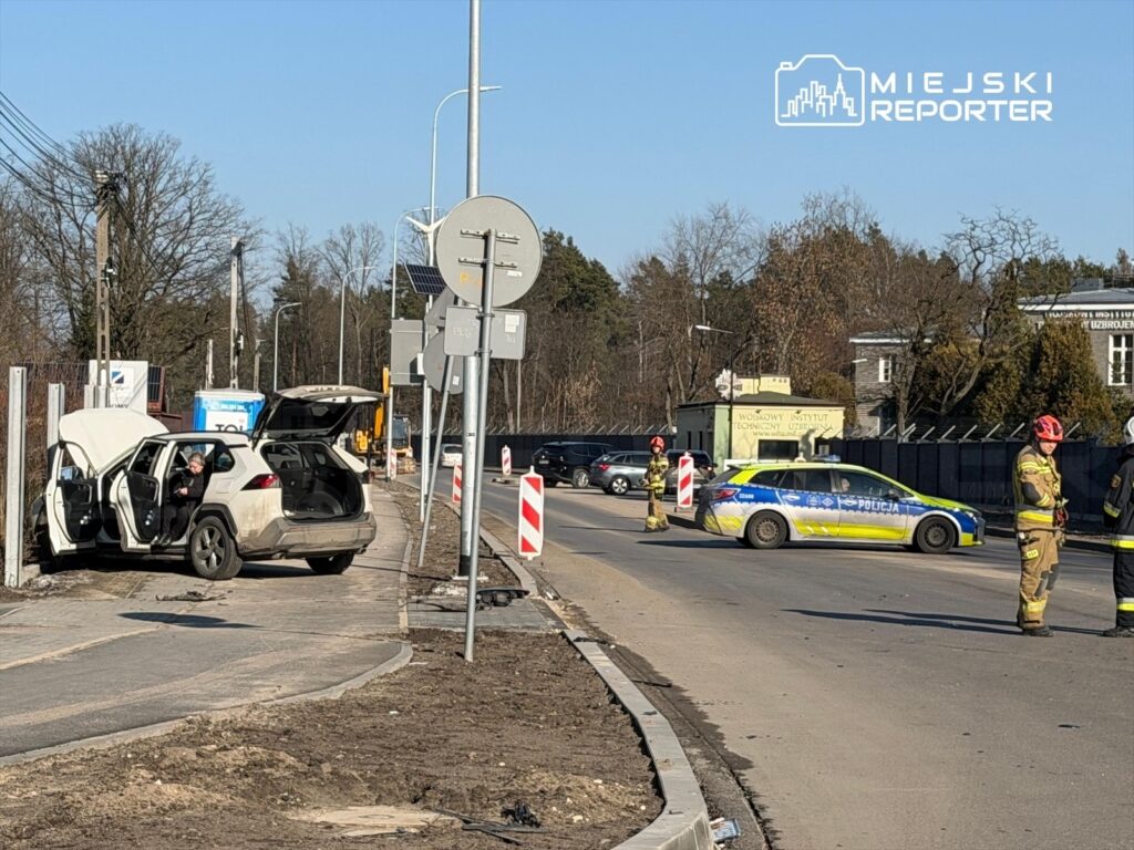 Kobieta siedząca w samochodzie z otwartą klapą bagażnika obok policyjnego radiowozu na drodze, w tle budynki i drzewa.
