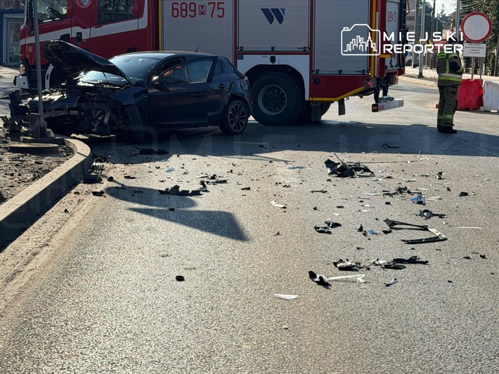 Czarny samochód osobowy z uszkodzonym przodem stoi obok wozu strażackiego, a wokół leżą fragmenty rozbitego pojazdu.